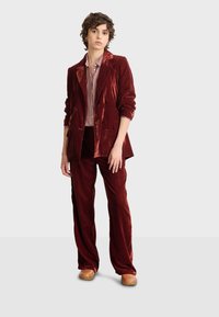 Sukienka burgundowa z weluru, składająca się z podwójnie zapiętej marynarki oraz szerokich spodni, w połączeniu z koszulą w kratę. Stylizację dopełniają klapki.