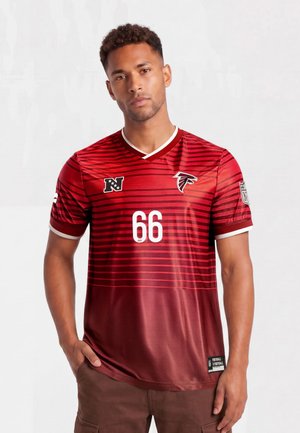 Homme portant un maillot rouge des Atlanta Falcons NFL avec le numéro 66 et un pantalon marron, debout avec une main dans la poche sur fond blanc.