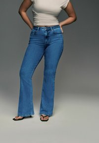 Uitlopende blauwe denim jeans met een hoge taille en een klassieke vijf-pocket ontwerp. G gedragen met zwarte flip-flops, in contrast met een aansluitende beige top.
