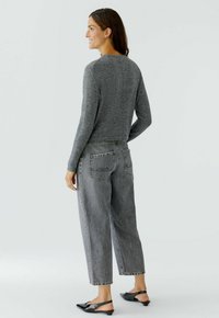 Eine Frau lächelt, trägt ein graues Langarmoberteil, hochgeschnittene lockere graue Jeans und schwarze spitz zulaufende Slingback-Ballerinas, während sie vor einem einfarbigen Hintergrund steht.
