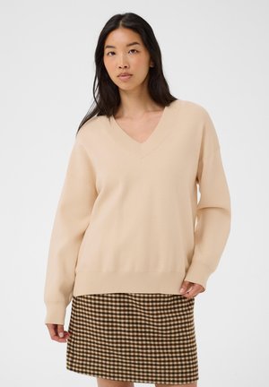 Jeune femme aux longs cheveux foncés portant un pull beige à col en V et une jupe marron à carreaux, debout sur un fond clair uni.