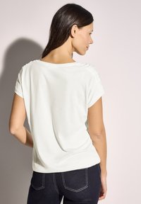 Camiseta de color claro, de corte holgado y mangas cortas, con detalles fruncidos en los hombros, combinada con jeans de mezclilla oscura.