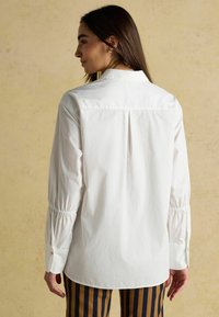 Weißes Hemd mit klassischem Kragen, gerafften Ärmeln und einem Rückenfalte, kombiniert mit vertikal gestreiften Hosen.