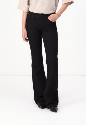 Fekete bővülő farmernadrág, amely denim anyagból készült, szűk derékrésszel, elülső zsebekkel, övhurkokkal és varrási részletekkel. Fekete hegyes cipővel viselve.