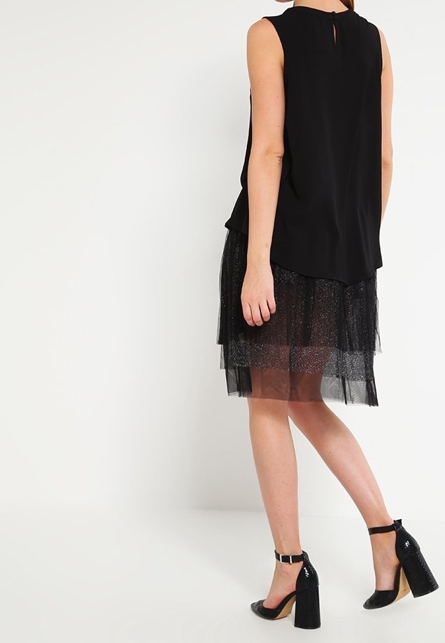 Top senza maniche nero con dettaglio a foro sul retro, abbinato a una gonna in tulle nero a strati con una leggera lucentezza. Scarpe con tacco nere.