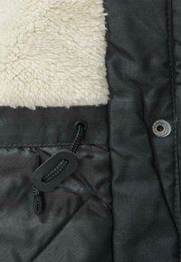 Svart quilted jakke med kremfarget saueskinnskrage. Har en svart snor og en trykknappåpning med detalj i sølv.