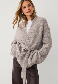 Frau in einem grob gestrickten beigen Cardigan mit gebundenem Gürtel über einem weißen Oberteil und dunkelbraunen Lederhosen, posierend vor einem schlichten Hintergrund.