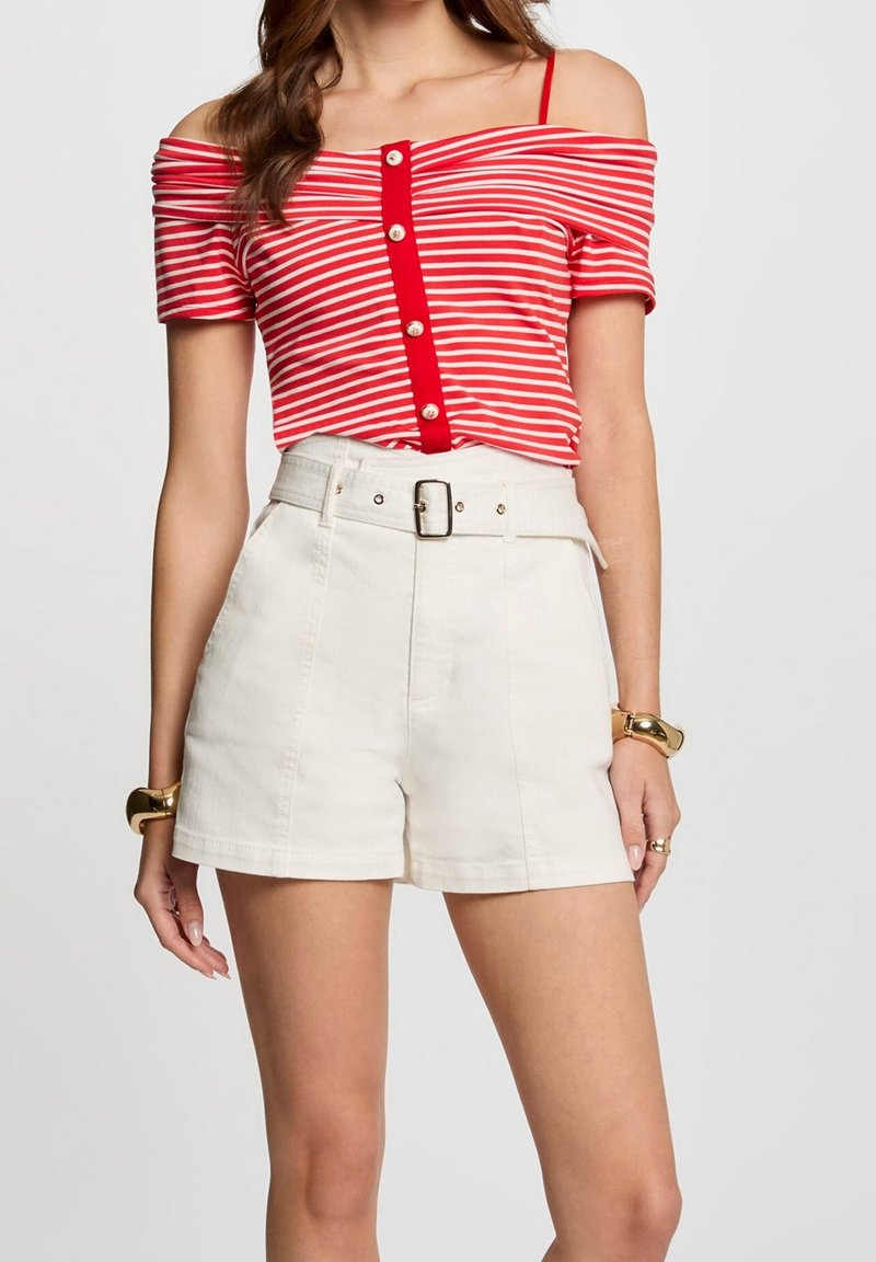 Femme portant un top rayé rouge et blanc épaules dénudées avec des boutons et un short blanc taille haute ceinturé, bracelets dorés aux deux poignets.