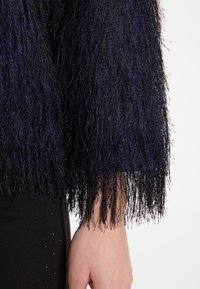 Schwarzes, fransiges Kleidungsstück mit dunklen blauen schimmernden Fäden. Verfügt über lange, strukturierte Ärmel und einen eng anliegenden schwarzen Saum mit subtilen Verzierungen.
