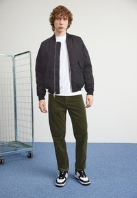 Giacca bomber nera, t-shirt bianca, pantaloni in velluto a coste verde oliva e sneakers bianco e nere; in piedi di fronte a un appendiabiti in filo.