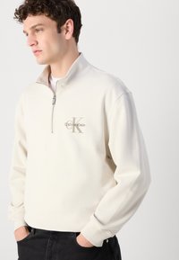 Felpa con zip di colore beige chiaro realizzata in tessuto morbido, con polsini a coste e un piccolo logo sul petto di un beige più scuro.