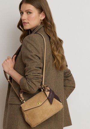 Vrouw met lang golvend haar draagt een bruin geruit blazer en draagt een lichtbruine suède handtas met een donker sjaaltje met patroon vastgeknoopt aan het hengsel.