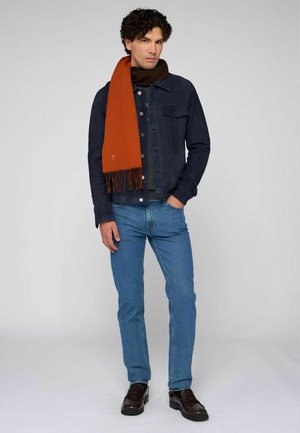 Marineblauer Wildlederjacke über einem grauen Pullover, hellblaue Jeans und ein braun-orange gestreifter Schal mit Fransen. Braune Loafers vervollständigen den Look.