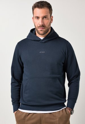 Mand med kort hår og skæg iført en marineblå JP1880 hættetrøje, hvid undertrøje, brune bukser og armbåndsur, stående neutralt mod en ensfarvet baggrund.