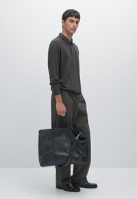 Schwarze Lederhandtasche mit zwei Griffen und einem abnehmbaren Schultergurt, getragen von einer Person in einem dunklen Pullover und gemusterten Hosen.