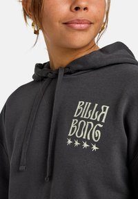 Sudadera gris con cordón, que presenta un logo "BILLABONG" de color claro y gráficos de estrellas. Textura suave, ajuste casual.