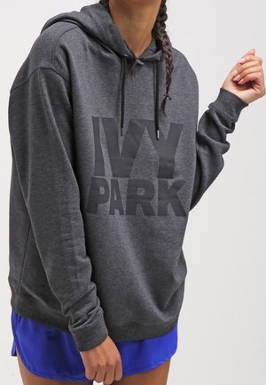 Person iført en mørkegrå IVY PARK-hoodie og lyseblå shorts med flettet hår mod en ensfarvet lys baggrund.