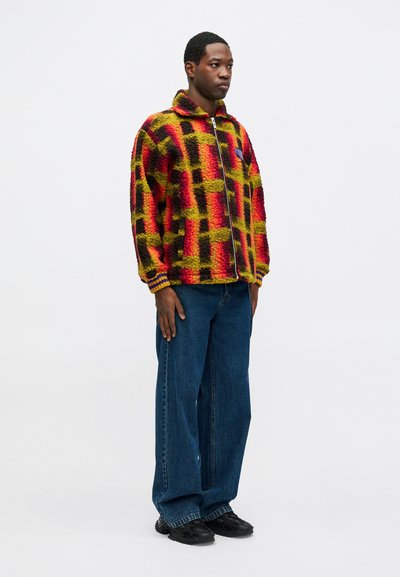 Raudonas, geltonas ir juodas languotas pūkinis švarkas su užtrauktuku, derinamas su laisvais mėlynais džinsais ir juodais batais.