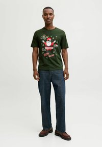 Camiseta verde de Navidad con un gráfico de Santa Claus, acentos rojos y texto festivo blanco. Usada con jeans azul oscuro y zapatos marrones.