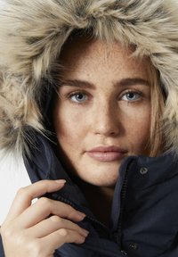 Námořnická modrá zimní bunda s kapucí zdobenou umělou kožešinou, se zapínáním na zip a texturovanou látkou. Modelova ruka drží zip.