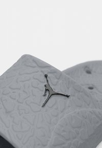 Sive teksturirane klizne sandale s oblikovanim dizajnom, koje imaju crni Jumpman logo na vrhu i ventilacijske rupe za prozračnost.