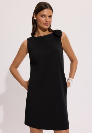 Kobieta w czarnej sukience bez rękawów z kokardą na ramieniu, nosząca perłowe kolczyki i perłową bransoletkę, stoi z rękami w kieszeniach.