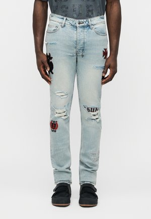 Ανοιχτό μπλε τζιν με φθορές, μπάλες και κείμενο, φορεμένα με μαύρα αθλητικά παπούτσια, εμφανιζόμενα από τη μέση μέχρι τα πόδια πάνω σε μονόχρωμο φόντο.