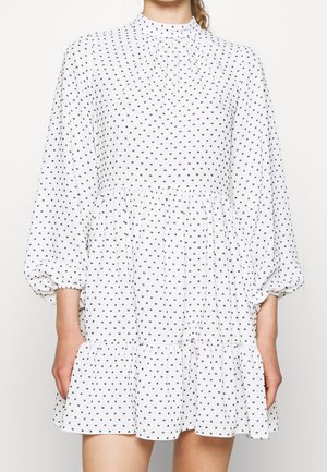 Vrouw die een witte jurk met lange mouwen draagt, met kleine zwarte vierkante patronen, een getailleerde taille en een geplooide zoom.