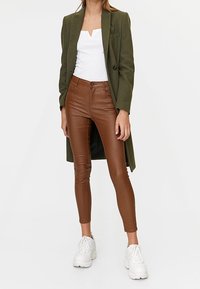 Olijfgroen lange jas, witte V-hals top, bruine strakke broek en witte dikke sneakers. Glad textuur en een op maat gemaakte, hedendaagse pasvorm.