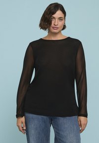 Schwarzes Langarmtop mit durchsichtigen Mesh-Ärmeln und Rundhalsausschnitt, kombiniert mit lockeren blauen Denim-Jeans. Hintergrund in sanftem Blau.