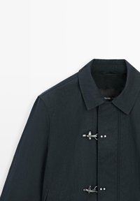 Cappotto blu scuro con colletto classico e due chiusure metalliche a forma di aeroplano sul davanti.