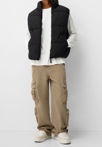 Svart vadderad väst med hög krage, bärs över en vit långärmad skjorta. Beige cargobyxor med sidofickor och ljusa sneakers.