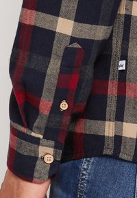 Granatowa koszula flanelowa w kratę z czerwonymi i beżowymi kwadratami, drewnianymi guzikami oraz wzorem jodełki na mankietach i w okolicy kieszeni.