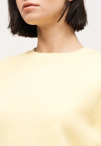 Femme aux cheveux courts et foncés portant un t-shirt jaune pâle à col rond, montrant le bas du visage, le cou et le haut du torse sur un fond uni.