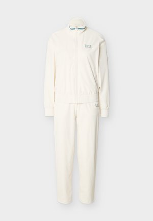 Λευκή ολόσωμη φόρμα με φερμουάρ και ελαστικά ρεβέρ στα μανίκια και τη μέση, με το λογότυπο EA7 στο στήθος και στο μηρό, εμφανίζεται σε απλό φόντο.