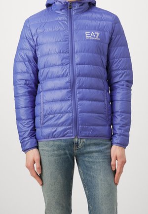 Veste matelassée violette avec fermeture éclair à l’avant, capuche et logo blanc EA7 Emporio Armani sur la poitrine, portée avec un jean bleu.