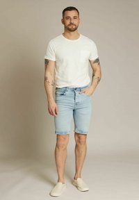 T-shirt blanche à manches courtes, shorts en denim bleu clair et chaussures slip-on beiges. Présente une poche sur la poitrine et des bras tatoués. Design simple.
