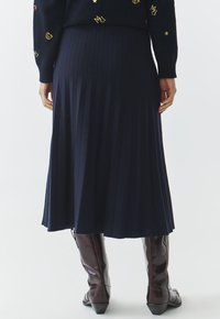 Marineblauwe geplooide midi-rok van jersey stof, met een gladde textuur, een licht uitlopende onderkant en gecombineerd met hoge, bruine leren laarzen.