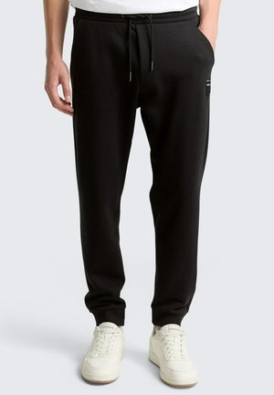 Mann trägt schwarze, schmal geschnittene Jogginghose mit Kordelzug an der Taille und weiße Sneaker, steht mit den Händen in den Taschen.