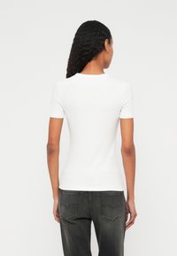 Biele priliehavé tričko s krátkym rukávom a okrúhlym výstrihom; rebrovaná textúra; nosené s tmavosivými džínsami.