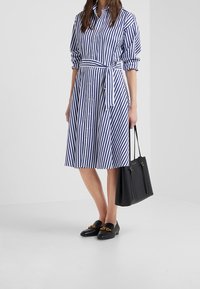 Kvinna i blå och vit randig klänning med knappar och midjeband, svarta loafers med gulddetalj, och bär en svart handväska.