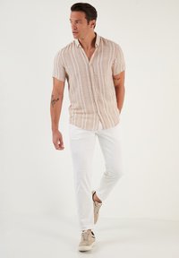 Camisa de manga corta para hombre, color beige claro con rayas verticales blancas, corte relajado, combinada con pantalones blancos y zapatillas ligeras.
