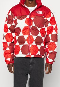 Röd och vit puffjacka med geometriskt mönster med trianglar och präglade mönster. Dragkedja fram, elastiska ärmslut och hög krage.