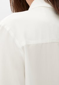 Biała koszula z tradycyjnym kołnierzykiem, miękką fakturą i gładkim wykończeniem, wyeksponowana z tyłu, podkreślająca detale szycia.