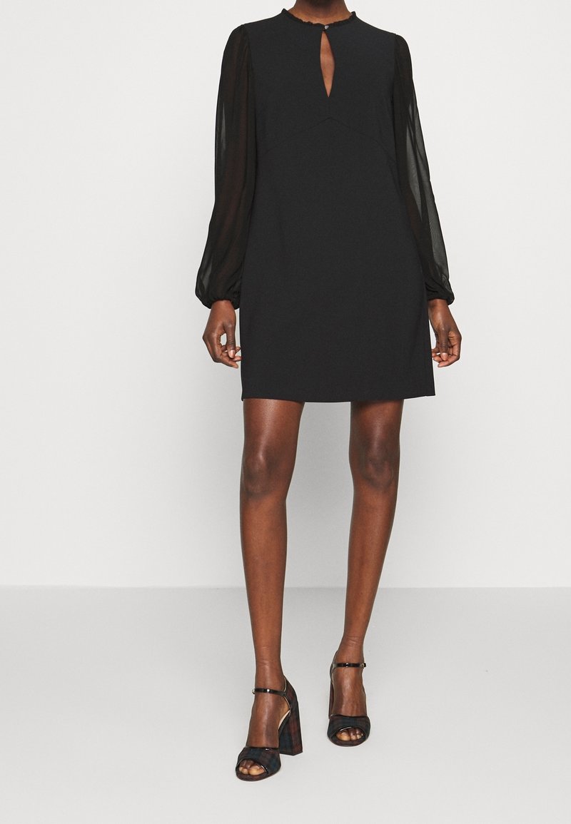 Femme portant une mini-robe noire à manches longues transparentes, un décolleté en trou de serrure à l'avant et des sandales noires à talons hauts avec bride à la cheville.