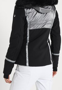 Schwarze Jacke mit silbernen Akzenten; verfügt über gesteppte Einsätze und eine Kapuze mit Kunstfell. Das Design umfasst schlanke Linien und eine taillierte Form.