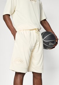 Lys cremefarvet athletic sæt, der inkluderer en tekstureret t-shirt med korte ærmer og matchende shorts med et prægede Lakers-logo. Inkluderer en sort basketball.