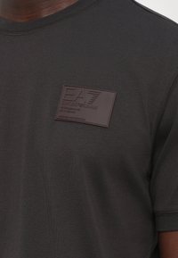 Mørk grå skjorte med en sydd brun rektangulær lapp med EA7 Emporio Armani tekst og merkeinformasjon på brystet.