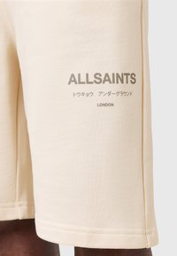 Shorts di cotone beige con la scritta "ALLSAINTS" e "LONDON" in stampa grigia, dalla texture liscia e design semplice.