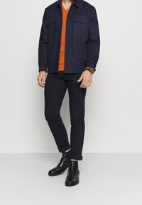 Mann in dunkelblauer Jacke mit Fronttaschen, orangefarbener V-Ausschnitt-Pullover, schwarzen hochgekrempelten Hosen, schwarzen Lederstiefeln und einer Armbanduhr am linken Handgelenk.